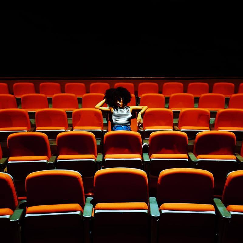Poltronas de uma sala de cinema, com destaque para uma moça sentada na poltrona do meio da imagem.