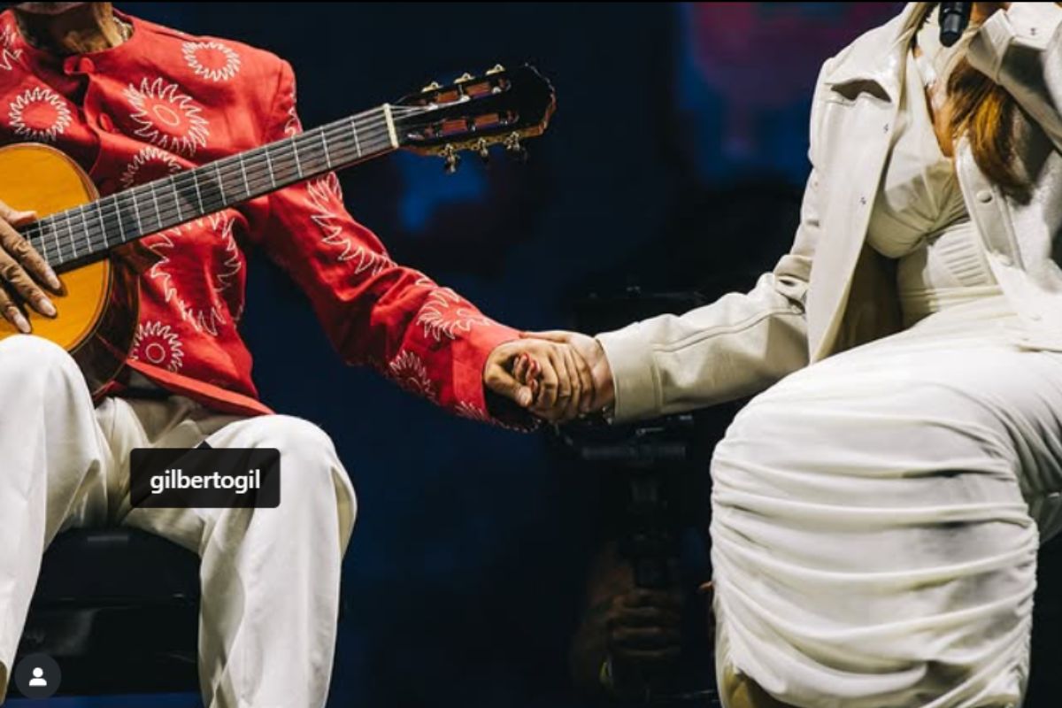 Close das mãos dadas de Preta Gil e Gilberto Gil durante show, ela vestida de branco e ele com violão